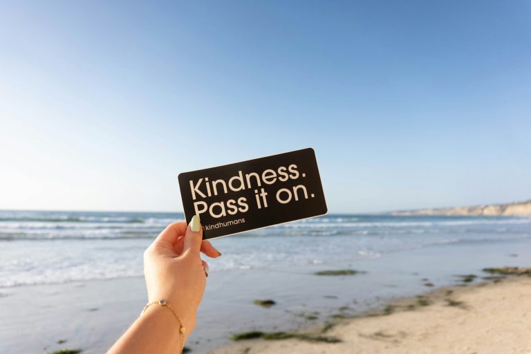 Hand holding a "Kindness: Pass it On" sticker in front of the ocean